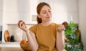 Vrouw twijfelt tussen een appel en een chocoladekoekje. De strijd tegen het verslaafd zijn aan eten is zichtbaar in haar blik.