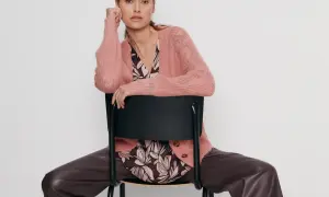 Elegancka kobieta w różowym swetrze Gerry Weber i brązowych spodniach z ekoskóry siedzi na czarnym krześle.