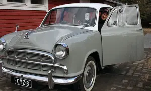 Man ler vid ratten i en grå Morris Oxford med öppen dörr.