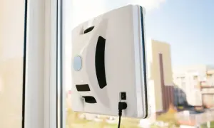 Biały robot do mycia okien przyczepiony do szyby, z widokiem na budynki w tle.