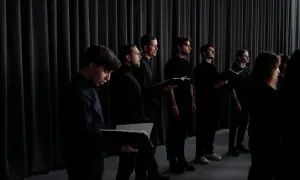 Chór męski w czarnych strojach, z nutami w rękach, przygotowuje się do wykonania utworu. Ich skupienie buduje napięcie, zapowiadając muzyczny **crescendo**.