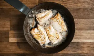 Quatre morceaux de lieu jaune poisson dorent dans une poêle en fonte sur un fond en bois.