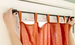 Dłonie montują czarny karnisz z pomarańczowymi zasłonami na drzwiach balkonowych.
