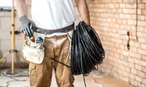 Elektryk z narzędziami w pasie trzyma zwój czarnego kabla. Czy będzie układany w ziemi w peszlu, czy bez?