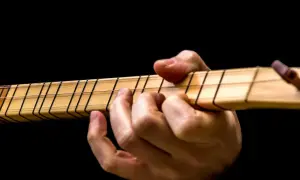 Dłoń gra na instrumencie, dociskając struny. Widoczne są progi i nazwy strun w gitarze.