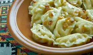 Pyszne pierogi ruskie z podsmażoną cebulką i skwarkami, podane w glinianej misce na haftowanej serwetce.