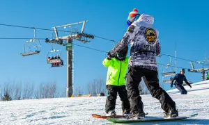 Instruktor snowboardingu uczy dziecko na stoku, w tle kolejka krzesełkowa.
