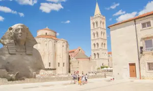 Zadar zabytki: kamienna sfinks, romańska rotunda i wieża dzwonnicza Katedry św. Anastazji. Turyści spacerują po placu.
