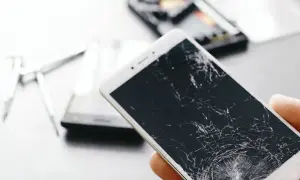 Dłoń trzyma telefon z pękniętym ekranem. Obok leżą narzędzia do naprawy.