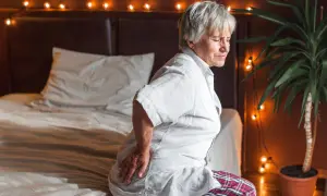 Elderly woman grimaces in pain, holding her hip. She looks like she slept on her hip wrong, sitting on a bed with festive lights.