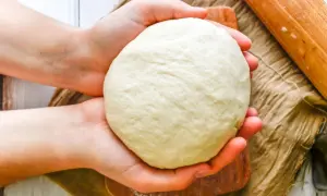 Dłonie trzymają kulę puszystego ciasta na pizzę włoską, gotowego do rozwałkowania. Obok wałek i lniana ściereczka.