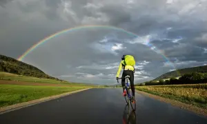 Rennradtour 3 Tage: Ein Radfahrer fährt unter einem Regenbogen auf nasser Straße.