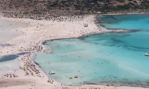 Słoneczna plaża na Krecie tętni życiem. Tłumy turystów kąpią się w turkusowej wodzie i odpoczywają na piasku pod parasolami.