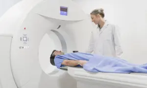 Pacjent w niebieskiej koszuli leży na stole tomografu. Lekarka w białym fartuchu nadzoruje badanie. Nie trzeba się rozbierać do tomografii.
