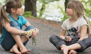 Dwie dziewczynki siedzą na ścieżce, rozmawiając. Jedna trzyma stokrotki. To inspiracja, jak nauczyć dziecko asertywności.