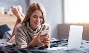 Uśmiechnięta dziewczyna leży na łóżku z telefonem w dłoniach, szukając informacji, jak doładować komuś konto w Play z karty na kartę. Obok leży laptop.