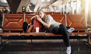 Zmęczona podróżniczka śpi na lotnisku, próbując pokonać jetlag. Obok niej plecak i kubek kawy.