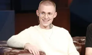 Młody mężczyzna z tatuażami na szyi, w okularach i swetrze, uśmiecha się. Mateusz Trąbka wiek nieznany, ale wygląda na młodego.