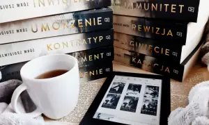 Stos książek Remigiusza Mroza, biały kubek z herbatą i czytnik Kindle.