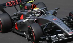 Un coche de **Audi F1** gris y negro con detalles rojos acelera en la pista. El piloto lleva casco amarillo y blanco.