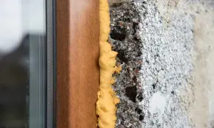Dettaglio di un telaio di finestra in legno con schiuma poliuretanica espansa che sigilla lo spazio tra il telaio e il muro grezzo. Questo mostra come modellare la schiuma poliuretanica per un isolamento efficace.