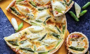 Herzhafte Quiche mit Spargel, goldbraun gebacken, auf einem orangen Tuch. Ein Stück ist angeschnitten, daneben liegen frische Spargelstücke.