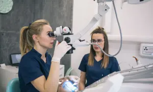 Dentystka z mikroskopem bada ząb pacjenta, by uniknąć chodzenia bez plomby. Pomaga jej asystentka.