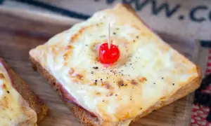 Ein klassisches Toast Hawaii mit geschmolzenem Käse, Schinken und einer Kirsche, serviert auf einem Holzbrett.
