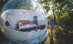 Une jeune femme admire sa **nuit bulle** confortable, un lit douillet dans une sphère transparente au milieu de la nature.