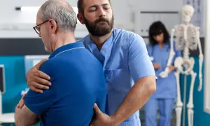 Fizjoterapeuta pomaga starszemu mężczyźnie z bólem pleców. W tle widać szkielet i drugą pielęgniarkę.
