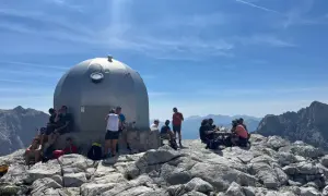 Gente descansa junto a un refugio en un viaje a Picos de Europa. El sol brilla en un cielo azul con nubes.