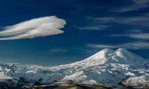Pokryty śniegiem Elbrus, najwyższy szczyt Rosji, góruje nad ośnieżonymi pasmami górskimi pod błękitnym niebem z charakterystyczną chmurą.