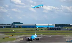 Dwa samoloty linii KLM na płycie lotniska i w powietrzu. Jeden kołuje, drugi wznosi się na tle chmur i budynków lotniska.