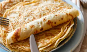 Ein Stapel goldbrauner Pfannkuchen, einer davon aufgerollt, liegt auf einem Teller. Ein Messer und eine Gabel liegen daneben. Perfekt für ein Pfannkuchen Rezept!