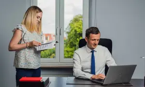 Kobieta z dokumentami i mężczyzna przy laptopie w biurze.