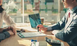 Osoba w garniturze stempluje dokument na biurku, obok laptopa i kalkulatora.