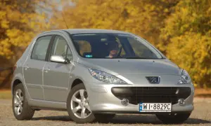 Srebrny Peugeot 307 z widocznymi żarówkami reflektorów, zaparkowany na tle jesiennych drzew.