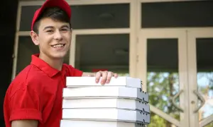 Uśmiechnięty pracownik młodociany w czerwonej koszulce i czapce niesie stos pudełek z pizzą.