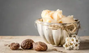 Naturalne masło shea na włosy w ozdobnej miseczce, obok orzechy shea i bukiecik kwiatów.