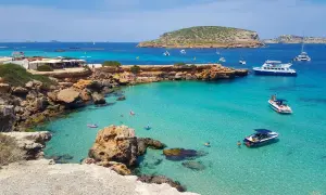 Plage idyllique à Ibiza avec eaux turquoise, rochers, bateaux et baigneurs. Parfait pour savoir où aller à Ibiza.