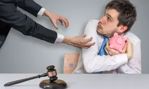 Młody mężczyzna kurczowo trzyma skarbonkę, gdy ręce w garniturze wyciągają się ku niemu. Młotek sędziowski leży na stole, symbolizując upadłość konsumencką.