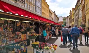 En livlig marknad i Prag med stånd fyllda av leksaker och blommor. Folk promenerar längs den soliga gatan.