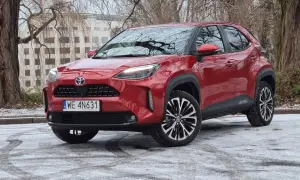 Czerwony Toyota Cross Hybrid na tle drzew i budynku. Auto stoi na ośnieżonej nawierzchni.
