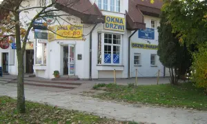 Budynek z żółtymi banerami "OKNA DRZWI" i "DRZWI PARAPETY", z drzewem po lewej.