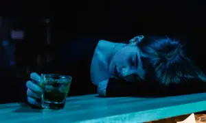 Zmęczona kobieta opiera głowę na blacie, obok szklanka z drinkiem. Mąż alkoholik obwinia mnie o wszystko, czuję się wyczerpana.