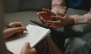 Dwie osoby w trakcie sesji terapeutycznej. Jedna notuje, druga gestykuluje, budując bliska relacja terapeutyczna.