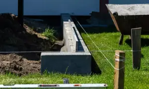 Budowa fundamentu pod ogrodzenie. Betonowe bloczki i poziomica wskazują precyzję prac. W tle taczka i kopiec ziemi.