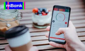 Dłoń trzyma smartfon z aplikacją mBank, na stole obok kawa i słoik z cukrem.