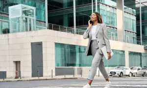 Kobieta w stylu business casual rozmawia przez telefon, niosąc laptopa. Idzie przez pasy przed nowoczesnym budynkiem.