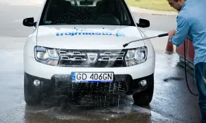 Mężczyzna myje białe Dacia Duster na myjni bezdotykowej, spryskując przód auta wodą pod ciśnieniem.